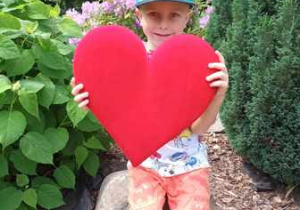 Natan w ogrodzie przedszkolnym pozuje do zdjęcia trzymając w dłoniach serce dla Mamy i Taty