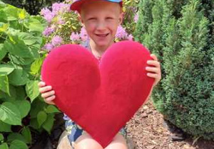 Maciek w ogrodzie przedszkolnym pozuje do zdjęcia trzymając w dłoniach serce dla Mamy i Taty