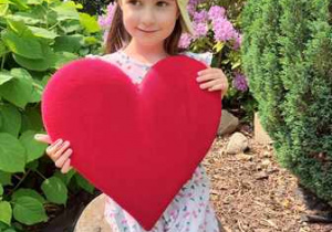 Nina w ogrodzie przedszkolnym pozuje do zdjęcia trzymając w dłoniach serce dla Mamy i Taty