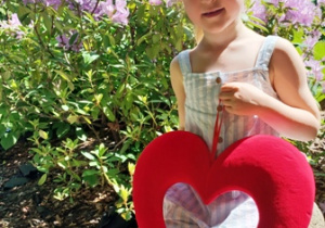 Heleneka w ogrodzie przedszkolnym pozuje do zdjęcia trzymając w dłoniach serce dla Mamy i Taty
