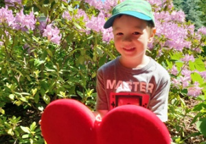 Wojtuś w ogrodzie przedszkolnym pozuje do zdjęcia trzymając w dłoniach serce dla Mamy i Taty