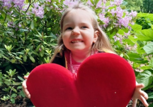 Emilka w ogrodzie przedszkolnym pozuje do zdjęcia trzymając w dłoniach serce dla Mamy i Taty