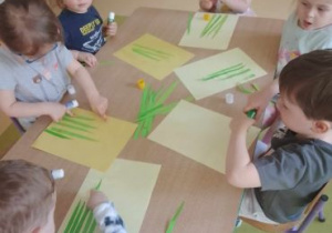 Dzieci siedzą przy stoliku i przyklejają trawę wyciętą z papieru
