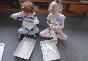 Dziewczynka i chłopiec siedzą na dywanie i wycinają paski z białego papieru