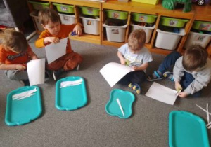 Dzieci siedzą w kręgu na dywanie i wycinają paski z białego papieru
