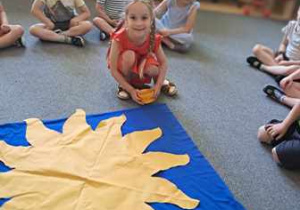 Dzieci siedzą w kręgu na dywanie, Anielka rozkłada talerzyki na promieniach "Urodzinowego Słoneczka"