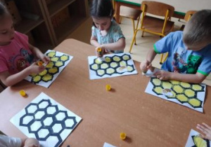 Dzieci siedzą przy stoliku i stemplują plastry miodu gąbką umoczoną w farbie