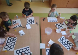 Dzieci siedzą przy stolikach i przyklejają kawałki papieru tworząc plaster miodu