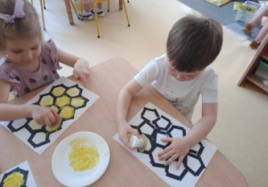 Dzieci siedzą przy stoliku i stemplują żółtą farbą