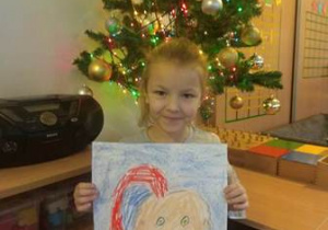 Dziewczynka stoi przy choince i prezentuje swój rysunek Świętego Mikołaja wykonany przy użyciu pasteli olejnych.