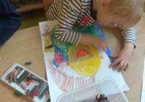 Dziewczynka przy stoliku rysuje portret Świętego Mikołaja pastelami olejnymi.