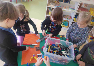 Dzieci stoją przy stole i budują z klocków znajdujących się w dużym pudełku