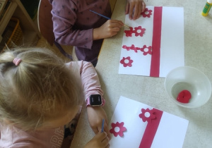 Dziewczynki malują farbami biało - czerwone kwiaty.