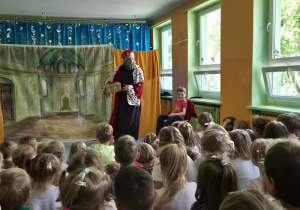 Chłopiec siedzi na krześle ustawionym z prawej strony przy oknie, a obok niego stoi aktor w przebraniu króla i trzyma w rękach złotą skrzynię