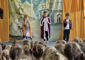 Dwie dziewczynki i jeden chłopiec stoją na scenie w przebraniach: Indianki, królowej i kowboja