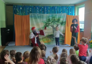 Dziewczynka stoi na środku sceny, obok niej stoi aktor w przebraniu króla oraz aktorka w zielonej długiej sukni
