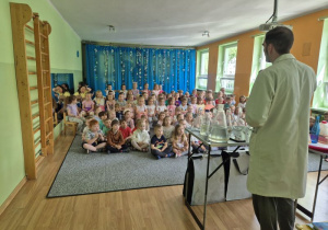 Dzieci siedzą i czekają na rozpoczęcie zabaw eksperymentalnych, na pierwszym planie stoi mężczyzna ubrany w biały fartuch, przed nim stoi stół