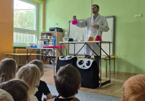 Mężczyzna ubrany w biały fartuch trzyma w ręku naczynie z różowym płynem, przed nim na stole stoją szklane kolby wypełnione kolorowymi cieczami