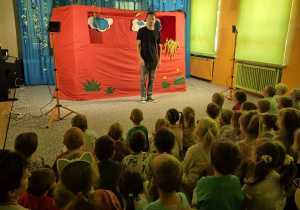 Dzieci oglądają przedstawienie siedząc na widowni, na scenie stoi aktor, w tle scenografia