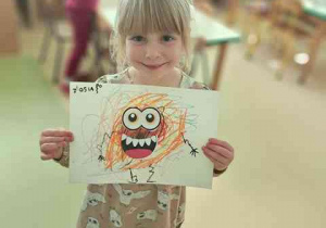 Dziewczynka trzyma w dłoniach wykonaną przez siebie pracę plastyczną pt. "Potworek pełen emocji"