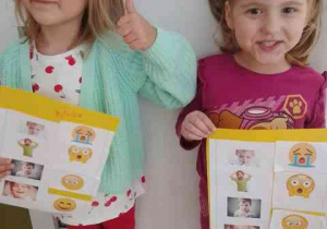 Liwia i Wiktoria z uśmiechem prezentują wykonane przez siebie karty pracy przedstawiające różne emocje