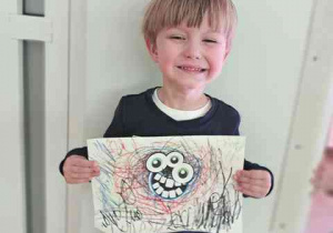 Chłopiec z uśmiechem pozuje do zdjęcia i trzyma w dłoniach wykonaną przez siebie pracę plastyczną pt. "Potworek pełen emocji"