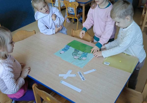 Dzieci siedzą przy stoliku i układają obrazek z rozsypanki oraz dobierają do niego podpis