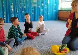 Prowadząca zajęcia trzyma w ręku marchewkę i pietruszkę oraz tłumaczy dzieciom znaczenie warzyw w diecie