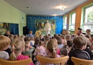 Dzieci siedzą na widowni i oglądają przedstawienie, na scenie stoi aktor w kostiumie zwierzątka