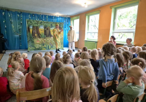 Dzieci siedzą na widowni i oglądają przedstawienie, na scenie stoją aktorzy w kostiumach zwierzątek