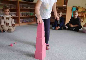 Dziewczynka układa różową wieżę, w tle widać dzieci siedzące w kręgu