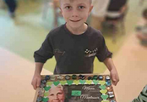 Natan trzyma w dłoniach utworzoną przez siebie kompozycję tworzącą ramkę do portretu Marii Montessori