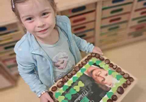 Dziewczyna trzyma w dłoniach wykonaną przez siebie pracę i wynosi na wystawę przez salą