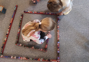 Dziewczynki układają szklane kamyczki na labiryncie z czerwonych sztang.