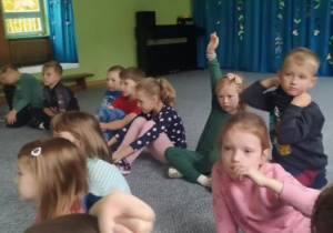 Dzieci siedzą w kręgu i słuchają prowadzącej zajęcia. Dziewczynka zgłasza się do odpowiedzi podnosząc rękę do góry