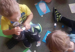 Dzieci siedzą na dywanie, przyklejają Mikołaja i prezenty na kartkę papieru