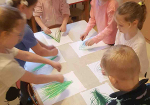 Dzieci stoją wokół stołu i rysują zielone kreski w rytm muzyki