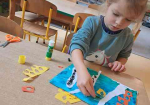 Dziewczynka siedzi przy stoliku i dokleja kawałki kolorowego papieru do swojej pracy plastycznej