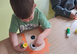 Chłopiec dokleja element papieru przedstawiający oko do swojej pracy plastycznej