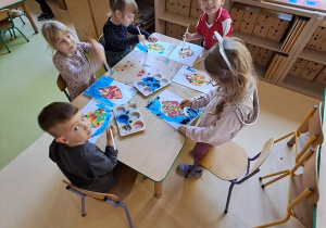 Dzieci w przy stoliku malują niebieską farbą tło swojej pracy plastycznej