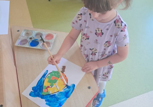Dziewczynka maluje niebieską farbą tło swojej pracy plastycznej
