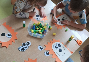 Dzieci przy stoliku w czasie podpisywania swojej pracy plastycznej przedstawiającej kompozycje jeża wykonaną z elementów kolorowego papieru