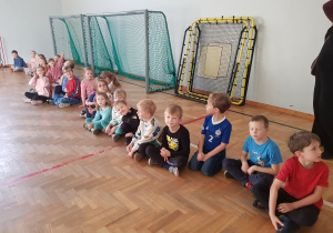 Dzieci siedzą na sali gimnastycznej w oczekiwaniu na zajęcia
