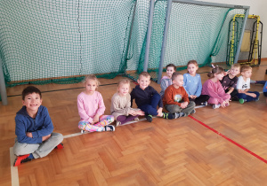 Dzieci siedzą na sali gimnastycznej w oczekiwaniu na zajęcia