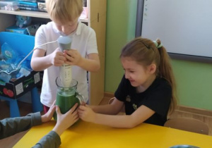 Dzieci siedzą przy stoliku, chłopiec używając blendera przygotowuje "Koktajl Shreka"