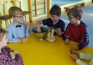 Dzieci siedzą przy stoliku, chłopiec wkłada banany do szklanego dzbanka