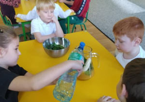 Dzieci siedzą przy stoliku, dziewczynka wlewa wodę do dzbanka z owocami i warzywami