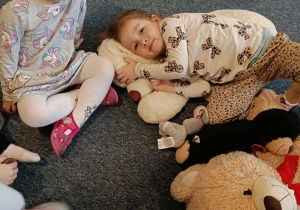 Dziewczynka siedzi na dywanie, druga dziewczynka leży wtulona w swojego misia