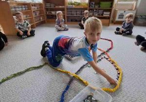 Chłopiec w czasie ćwiczenia ciszy dokłada szklane kamienie w zaznaczoną SIgumą przestrzeń na dywanie, w tle widać dzieci siedzące na dywanie