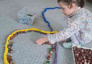 Dziewczynka dokłada kasztany i kamienie w zaznaczoną SIgumą przestrzeń na dywanie